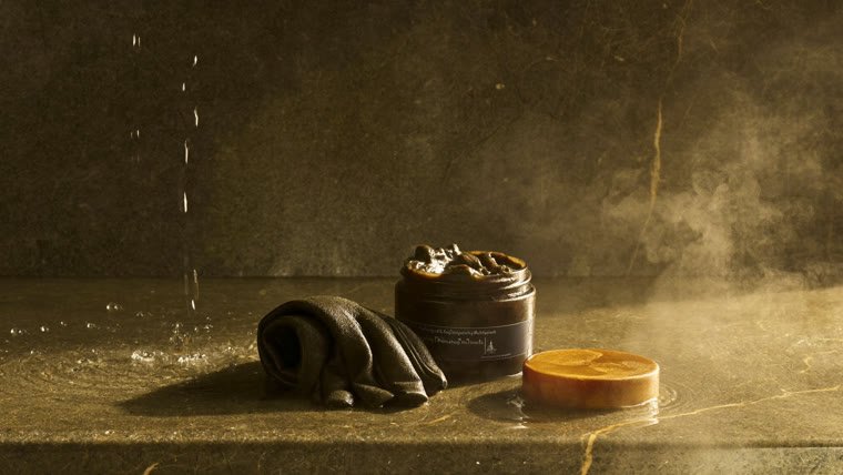 Pot de savon noir beldi ouvert et gant kessa plié sur la pierre humide d'un hammam, dans la vapeur