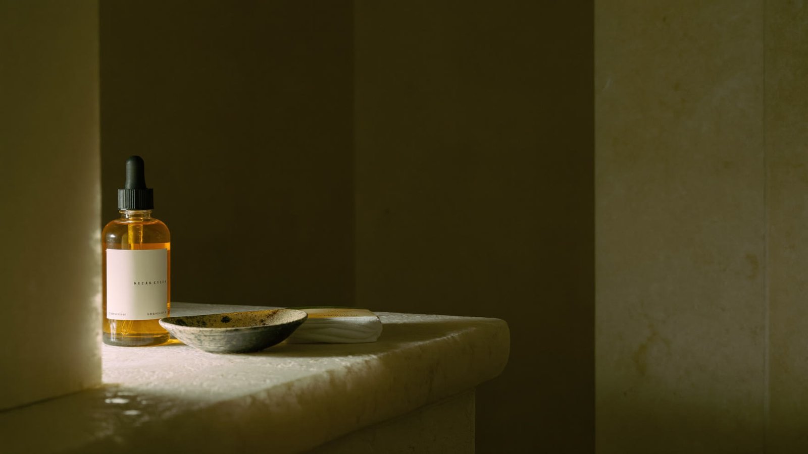 Coupelle en céramique, carré de coton et flacon d'huile d'argan sur une étagère de pierre, à la lumière chaude d'une lampe le soir