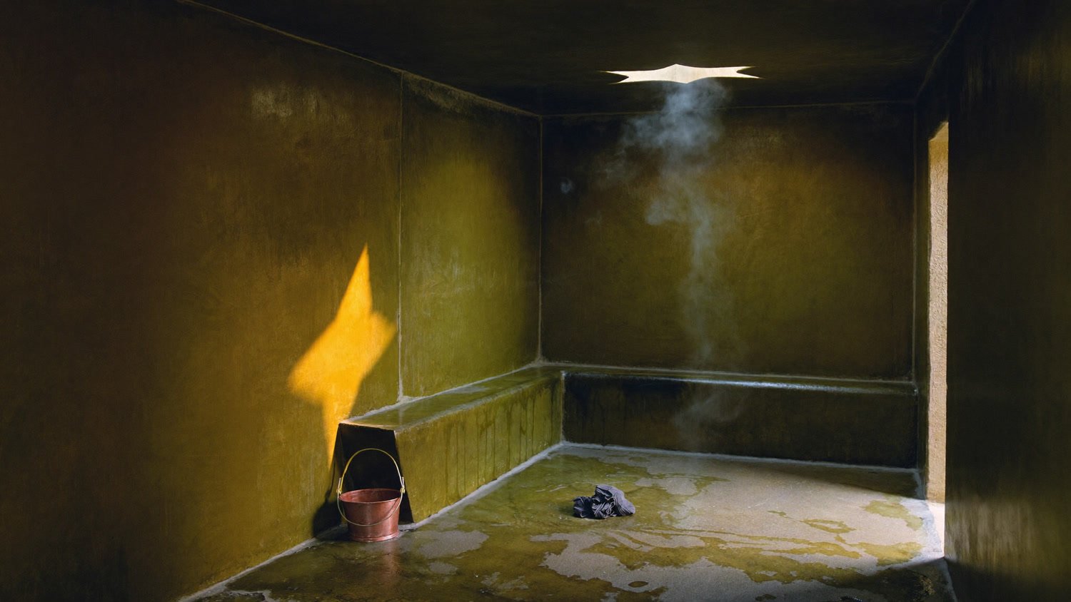 Intérieur d'un hammam marocain à l'aube, banc de pierre olive, seau en cuivre et vapeur traversée par un rayon de lumière
